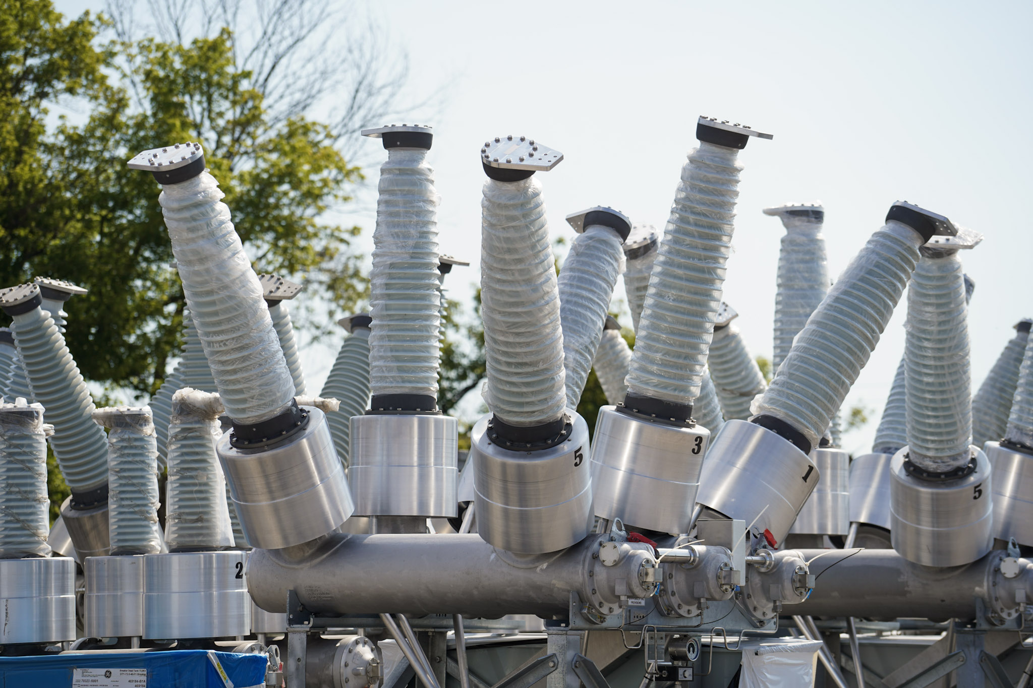 Dead-tank high-voltage breakers with wrapped bushings staged in the JCL yard against a blue sky